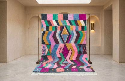 A vibrant, multicolored geometric patterned rug displayed on a wooden stand in a minimalist, warm beige interior room.