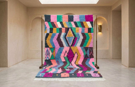 A vibrant, multicolored geometric patterned rug displayed on a wooden stand in a minimalist, warm beige interior room.