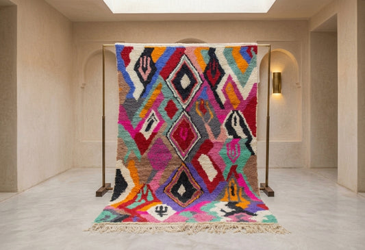 A vibrant Moroccan-style woven rug displayed on a metal stand in a minimalist beige hallway with arched doorways and a skylight.