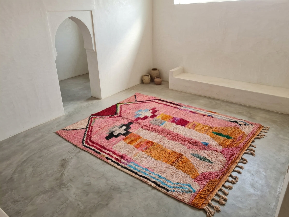 A colorful Moroccan-style woven area rug with geometric patterns and tassels, displayed on a concrete floor in a minimalist, off-white room with a built-in bench and arched doorway.