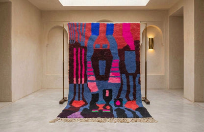 A large abstract patterned rug displayed on a metal stand in a minimalist beige gallery space. The rug features a bold, colorful design with vertical stripes and organic shapes in blue, pink, purple, black, and red, finished with a fringed edge.