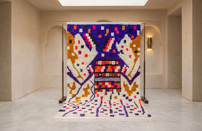 A large woven rug displayed on a metal stand in a minimalist gallery space. The rug features a highly colorful, abstract geometric pattern with purple, orange, white, and red elements.