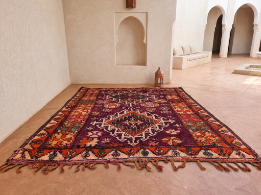 A large traditional Moroccan-style area rug with a complex geometric and floral pattern in deep purple, orange, red, and cream, laid out on a warm beige stone floor in a Moroccan courtyard.