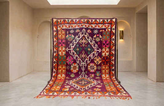 A large, ornate traditional rug with a deep purple and orange geometric pattern, displayed hanging from a black support frame in a minimalist beige room.