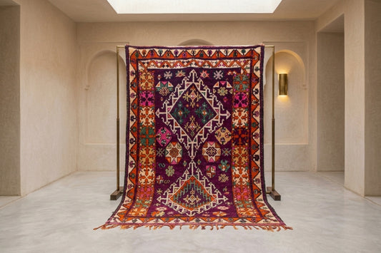 A large, intricately patterned Moroccan kilim rug hanging from a black metal stand in a minimalist, warm-toned gallery space with beige walls and arches.
