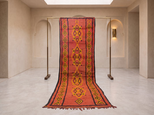 A long red and orange patterned wool runner rug with yellow and black geometric motifs, hanging from a metal stand in a minimalist beige room with arched walls and a skylight.
