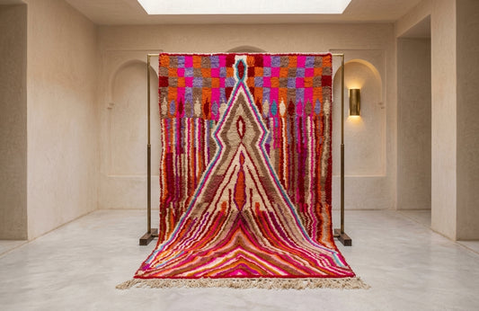 A large, vibrant Moroccan-style woven rug displayed on a black metal stand in a minimalist, warm-toned gallery room. The rug features a complex geometric pattern with a central diamond motif in shades of red, pink, orange, beige, and purple.