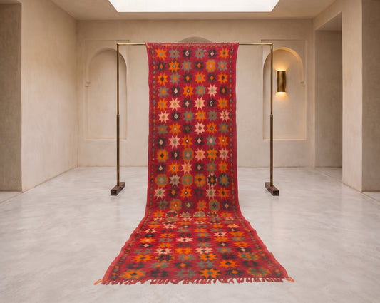 A long kilim area rug with a vibrant red border and a multicolor geometric star pattern, hanging from a metal stand in a minimalist, warm-toned gallery space.