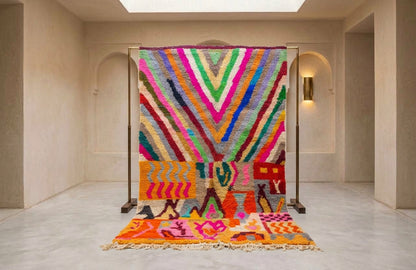 A large, vibrant, multicolored Moroccan-style woven rug displayed on a black metal stand in a minimalist, warm-toned gallery room.