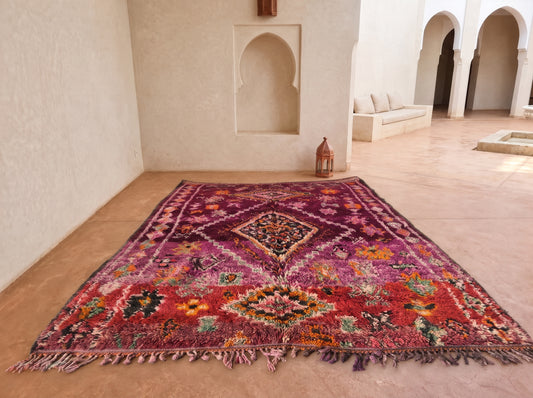 A large Moroccan-style area rug with a vibrant red, purple, and orange floral pattern and fringed edges, displayed on a terracotta tiled floor in a minimalist, sunlit courtyard with cream-colored walls and arches.
