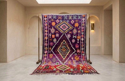 A large Moroccan-style wool rug with a complex geometric diamond pattern in purple, red, orange, and black, hanging from a black metal stand in a minimalist beige hallway.