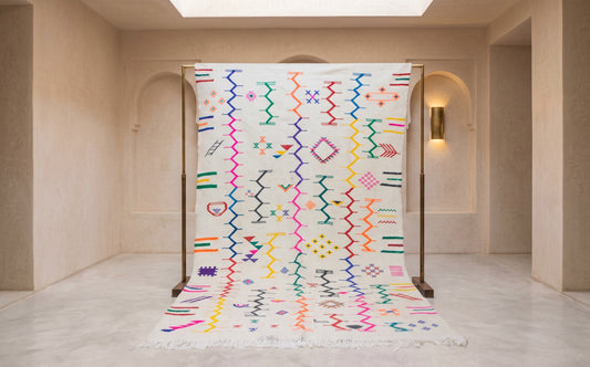 A large white Moroccan-style woven rug with vibrant multicolored geometric patterns, displayed on a wooden stand in a minimalist beige hallway.