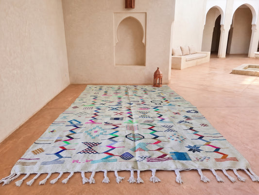 A large rectangular Moroccan-style rug with a cream base and colorful geometric patterns, laid out on a terracotta tiled courtyard floor with white tassels at the ends.