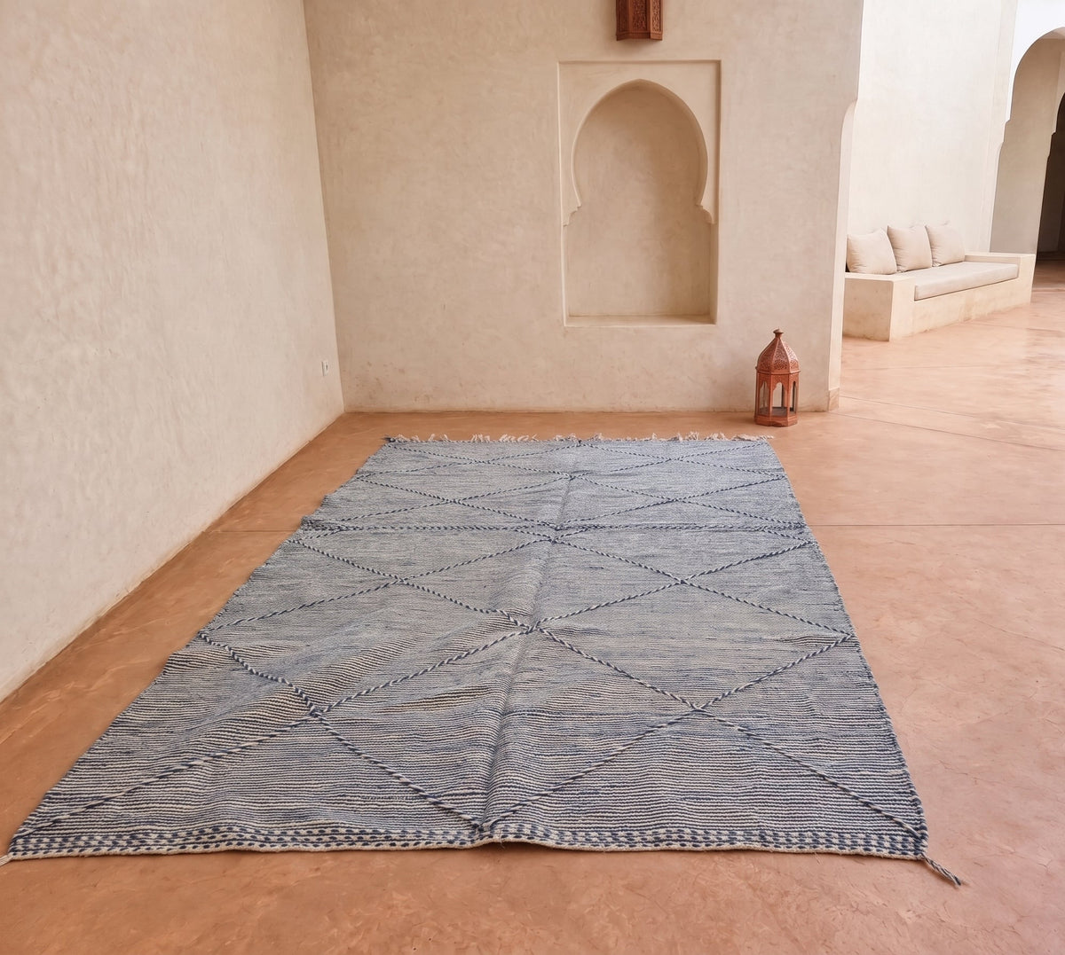 A large rectangular woven area rug with a blue and beige geometric diamond pattern, laid out on a tiled floor in a shop setting with other colorful textiles visible in the background.