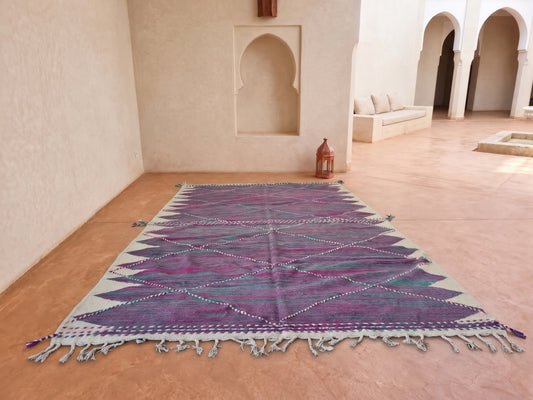 A rectangular Moroccan-style woven rug with a geometric diamond pattern in purple, teal, and pink, laid out on a terracotta tiled floor in a minimalist courtyard with beige walls and arches.