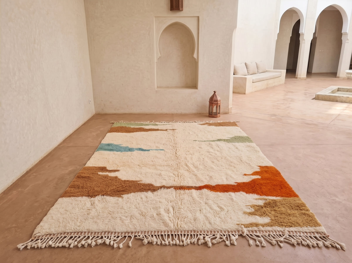 A rectangular Moroccan-style area rug with a textured, hand-tufted surface and tasseled fringe, displayed on a terracotta floor in a minimalist courtyard with arched alcoves.