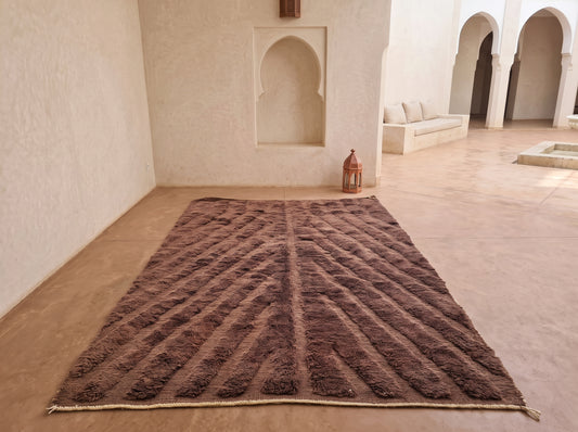 A large rectangular brown shag rug with a raised geometric chevron pattern, displayed on a terracotta-colored floor in a Moroccan-style courtyard with white walls and arches.