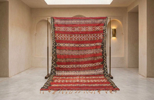 A large woven rug with red, beige, and black geometric patterns, displayed hanging from a black metal frame in a minimalist beige room.