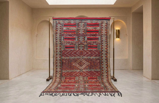 A large patterned area rug displayed hanging from a black metal stand in a minimalist beige interior room with arched walls and a skylight.