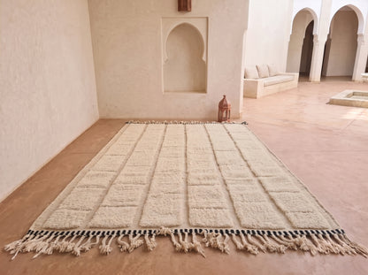 A large rectangular cream-colored rug with a grid pattern and tasseled fringe, laid out on a terracotta tiled floor in a Moroccan-style room with arched alcoves.