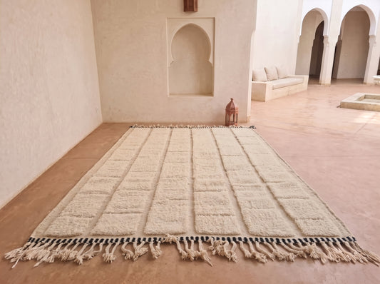 A large rectangular cream-colored rug with a grid pattern and tasseled fringe, laid out on a terracotta tiled floor in a Moroccan-style room with arched alcoves.