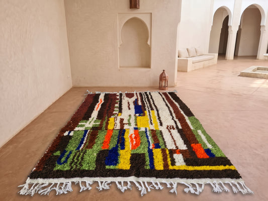 A colorful Moroccan-style woven rug with white fringe, displayed on a terracotta tiled floor in a minimalist, warm-toned Moroccan courtyard with arched alcoves.
