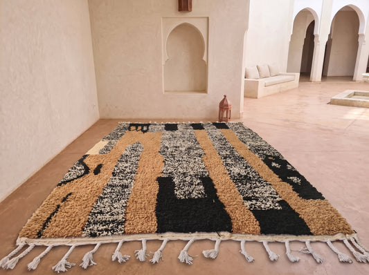 A large Moroccan-style area rug with a textured geometric pattern in black, tan, and cream, featuring white fringe tassels along the bottom edge, displayed on a terracotta tiled floor in a Moroccan-style room with arched alcoves.