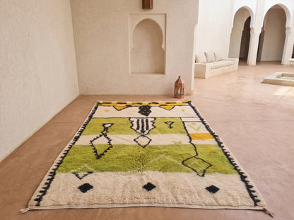 A rectangular handwoven rug with a Moroccan tribal design, featuring stripes of olive green, yellow, black, and cream, displayed on a terracotta tiled floor in a minimalist Moroccan courtyard.