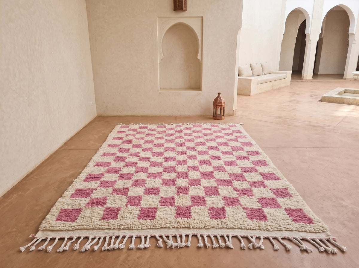 A rectangular woven rug with a pink and cream checkerboard pattern and tasseled edges, displayed on a terracotta floor in a Moroccan-style courtyard.