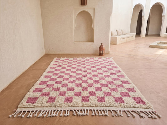 A rectangular woven rug with a pink and cream checkerboard pattern and tasseled edges, displayed on a terracotta floor in a Moroccan-style courtyard.