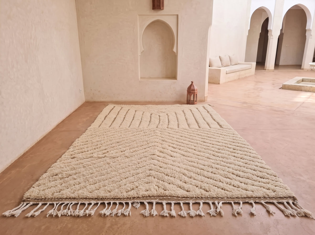 A large cream-colored area rug with a textured chevron pattern and fringe, displayed on a terracotta tiled floor in a Moroccan-style room with arched alcoves.