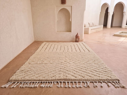A large cream-colored area rug with a textured chevron pattern and fringe, displayed on a terracotta tiled floor in a Moroccan-style room with arched alcoves.