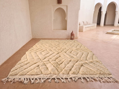 A large beige Moroccan-style rug with a raised geometric diamond pattern and tasseled fringe, displayed on a terracotta tiled floor in a minimalist, sunlit room with arched alcoves.