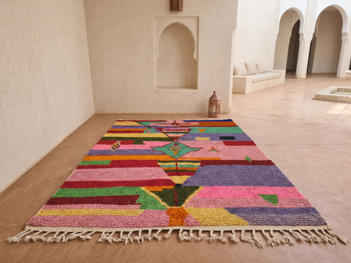A colorful Moroccan-style woven area rug with a geometric tree motif and cream fringe, displayed on a terracotta floor in a minimalist Moroccan courtyard.
