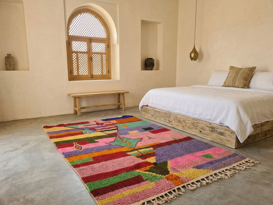 A colorful geometric area rug with fringe, styled in a rustic, warm-toned bedroom next to a wooden bed frame.