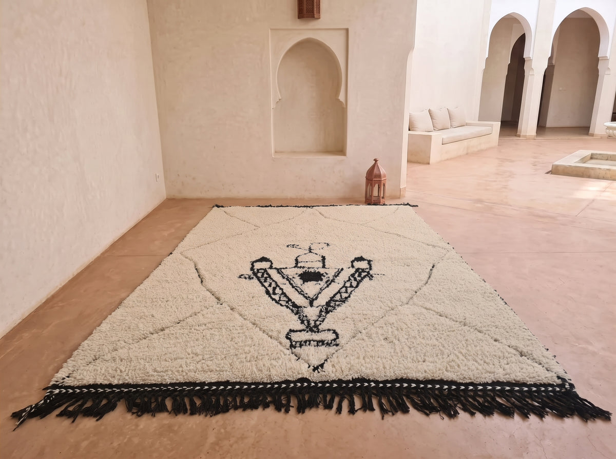 A large rectangular cream-colored rug with black fringe and a central black geometric emblem, displayed on a terracotta floor in a Moroccan-style room with arched alcoves.