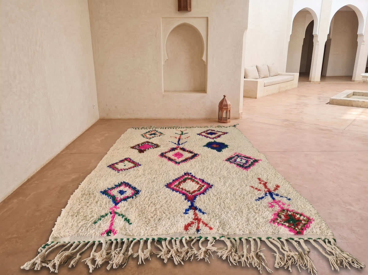 A rectangular Moroccan-style rug with a cream base and colorful geometric patterns, laid out on a terracotta tiled floor in a Moroccan courtyard with white stucco walls and arches.
