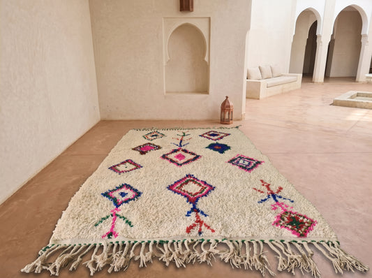 A rectangular Moroccan-style rug with a cream base and colorful geometric patterns, laid out on a terracotta tiled floor in a Moroccan courtyard with white stucco walls and arches.