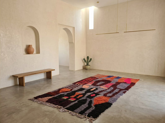 A large rectangular area rug with a bold abstract pattern in black, orange, pink, purple, and gray, styled on a concrete floor in a minimalist, warm-toned room with stucco walls and arches.