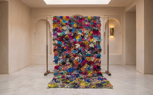 A large, vibrant, multicolored textured wall hanging or tapestry displayed on a minimalist gold metal frame in a modern, warm-toned gallery space.