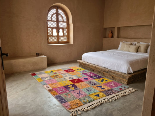 A colorful Moroccan-style patchwork area rug with fringe, displayed on a concrete floor in a rustic, earth-toned bedroom next to a wooden bed.