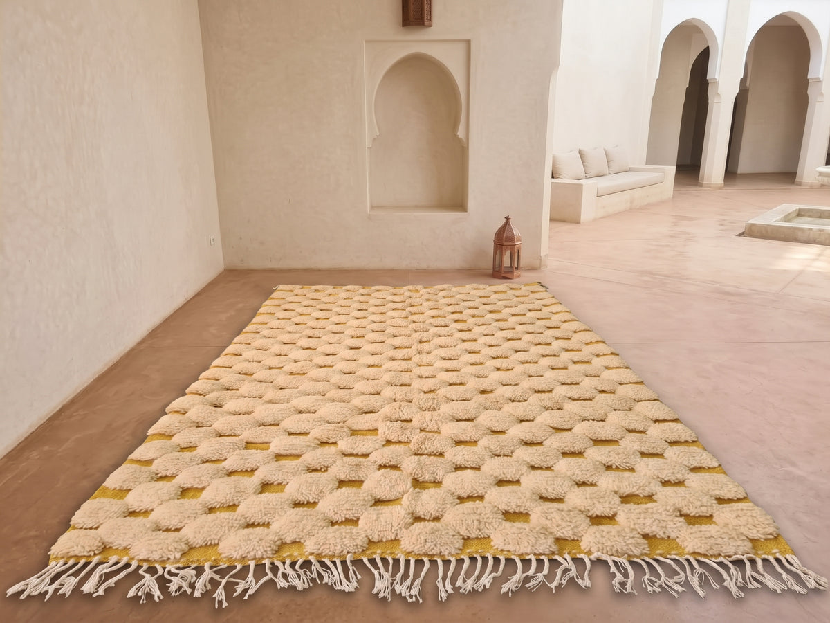 A rectangular Moroccan-style area rug with a textured geometric pattern in cream and mustard yellow, laid out on a terracotta tiled floor in a minimalist, sunlit room with arched alcoves.