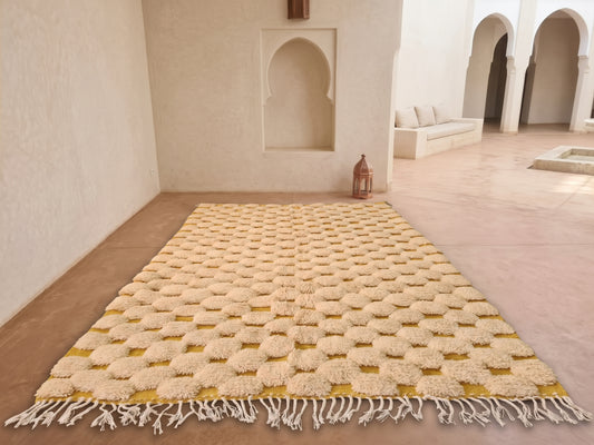 A rectangular Moroccan-style area rug with a textured geometric pattern in cream and mustard yellow, laid out on a terracotta tiled floor in a minimalist, sunlit room with arched alcoves.