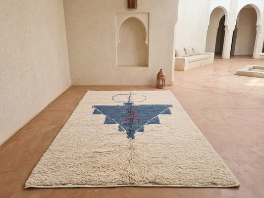 A large cream-colored shag rug with a central blue and red geometric pattern, displayed on a terracotta tiled floor in a Moroccan-style courtyard with white walls and arches.