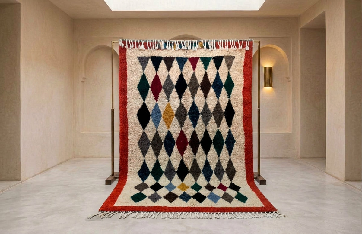 A large Moroccan-style woven rug with a geometric diamond pattern in multiple colors, hanging on a black metal stand in a minimalist beige hallway.