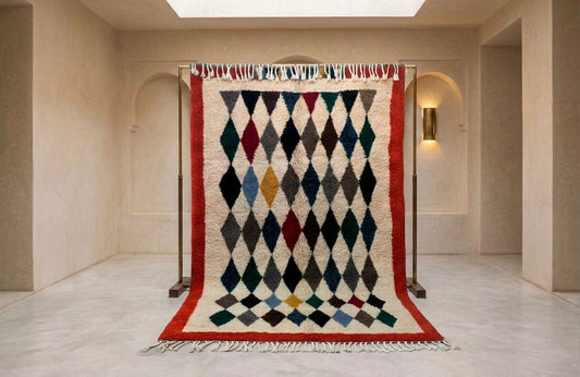 A large Moroccan-style woven rug with a geometric diamond pattern in multiple colors, hanging on a black metal stand in a minimalist beige hallway.