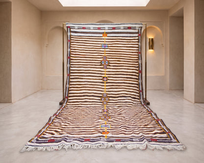 A large Moroccan-style wool rug displayed flat on a stand in a minimalist beige room. The rug features horizontal stripes in brown, beige, and white, with a central vertical band of orange, yellow, and blue geometric patterns, and white fringe at the bottom.