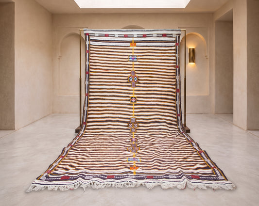 A large Moroccan-style wool rug displayed flat on a stand in a minimalist beige room. The rug features horizontal stripes in brown, beige, and white, with a central vertical band of orange, yellow, and blue geometric patterns, and white fringe at the bottom.