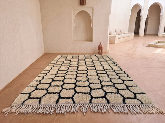 A large rectangular area rug with a black and cream geometric honeycomb pattern and tasseled fringe, displayed on a terracotta floor in a minimalist Moroccan-style room with arched alcoves.
