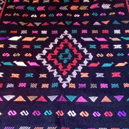 A rectangular black woven rug with a colorful geometric pattern, displayed on a shop floor alongside other rugs and textiles.
